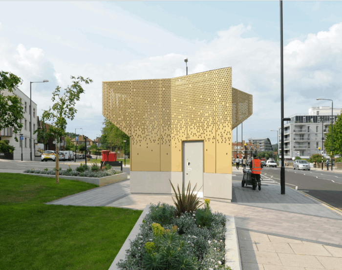 A striking gold, laser-cut perforated facade gives this public toilet block a landmark presence on the street corner."
Alt text: "A freestanding public toilet building clad in gold perforated metal panels with a diamond-pattern cutout design, sited on a paved corner with flower beds and grass, a street cleaner pushing a cart past it, and residential buildings and parked cars in the background